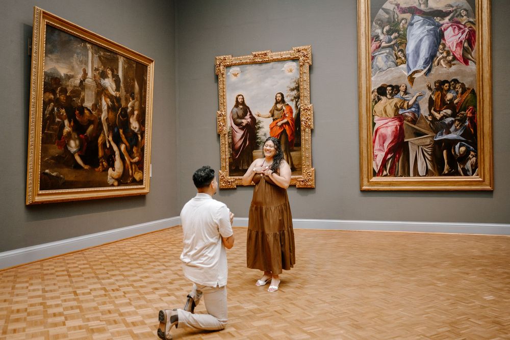 Man proposing on one knee to woman in art museum gallery surrounded by classical paintings.