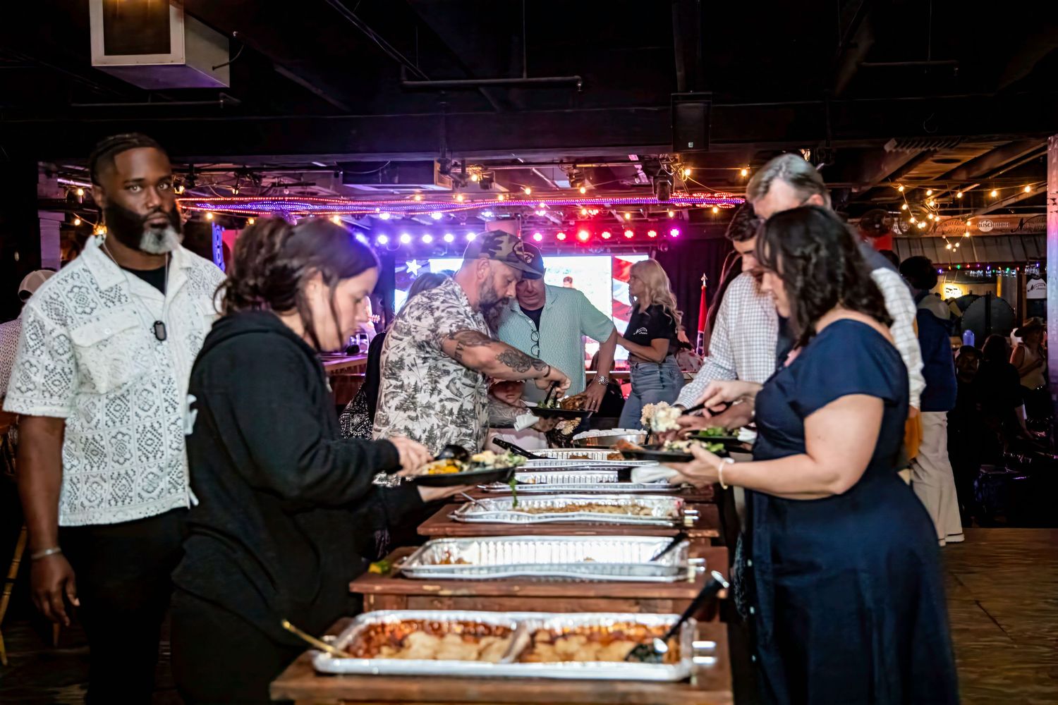 Guests serving themselves at a buffet dinner during an indoor event with colorful stage lighting in the background.