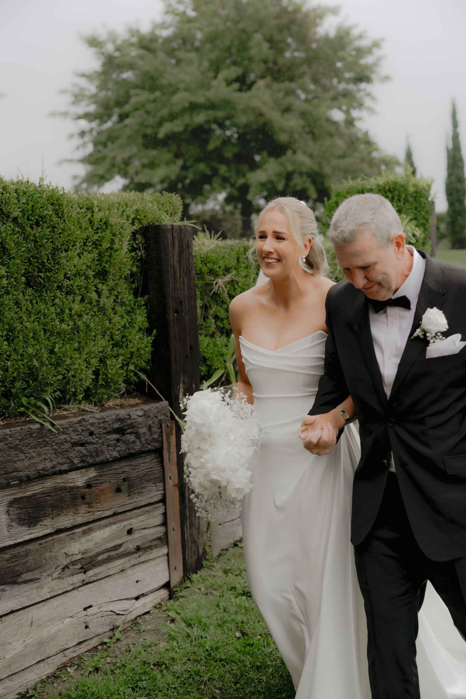 A bride in a white off-shoulder gown holds a bouquet while walking with father in a tuxedo down wedding aisle. Borrodell Vineyard Orange