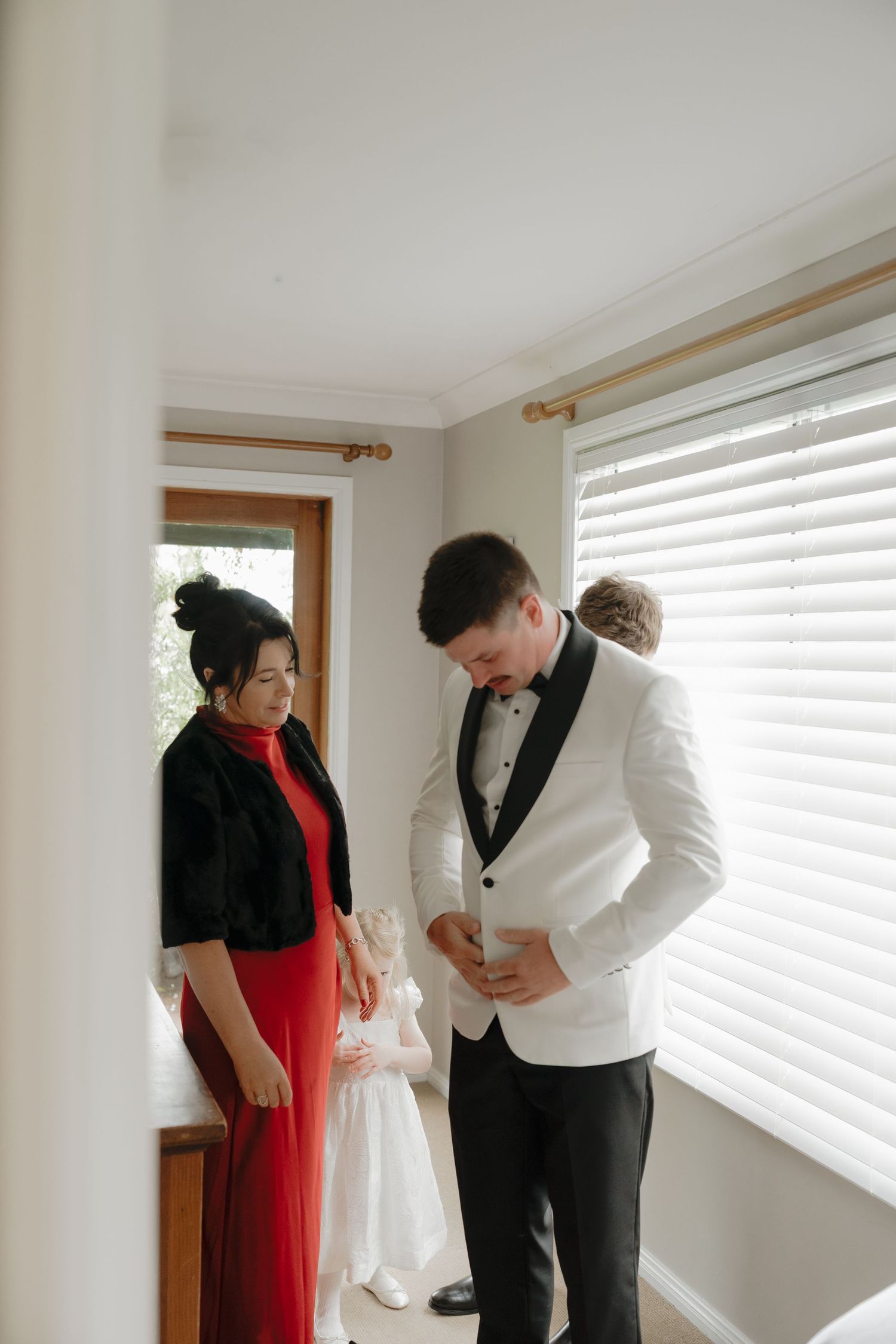 Groom in white tuxedo jacket adjusting his suit while a woman in red dress and black jacket stands nearby indoors. Borrodell Vineyard Orange