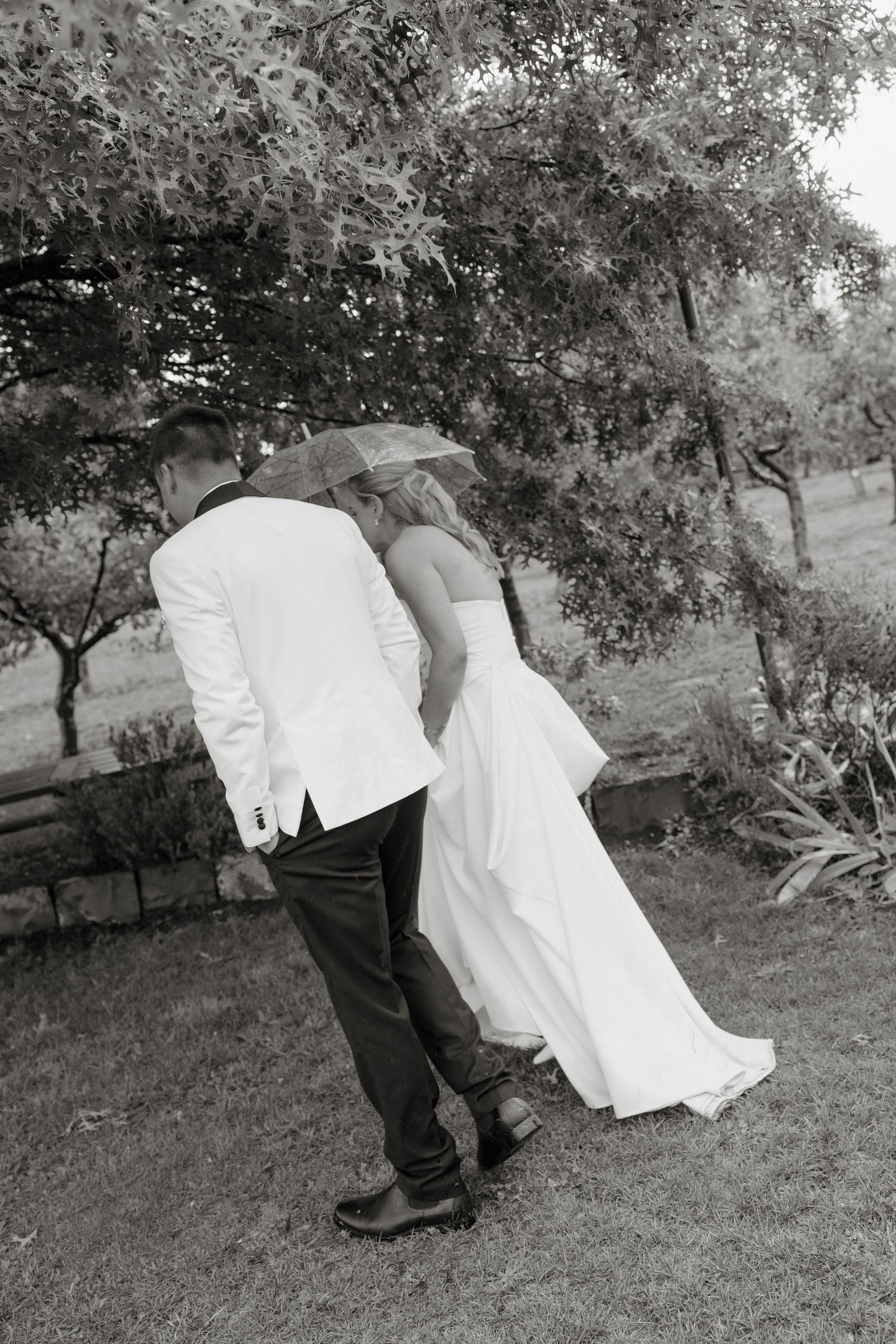 Black and white photo of a bride and groom walking outdoors under an umbrella on their wedding day. Borrodell Vineyard Orange