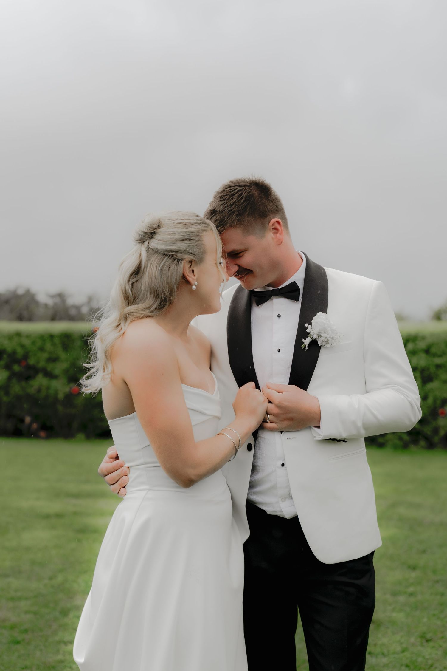 Bride and groom share an intimate moment outdoors, she in a white strapless gown, he in a white tuxedo with bow tie. Borrodell Vineyard