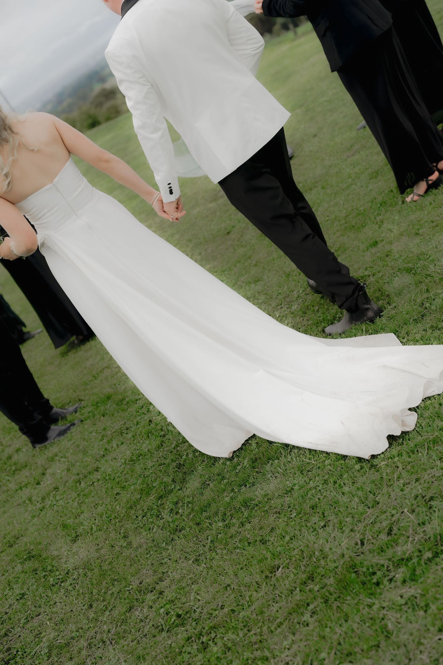 Bride in white gown with train holds groom's hand outdoors on green grass, surrounded by wedding party in black suits. Borrodell Vineyard