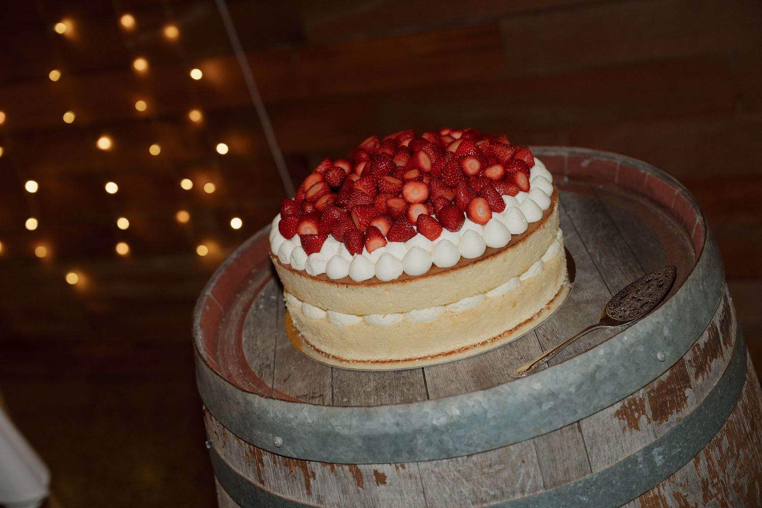 A strawberry topped white cream cake displayed on a rustic wooden barrel with warm bokeh lights in the background. Borrodell Vineyard Orange