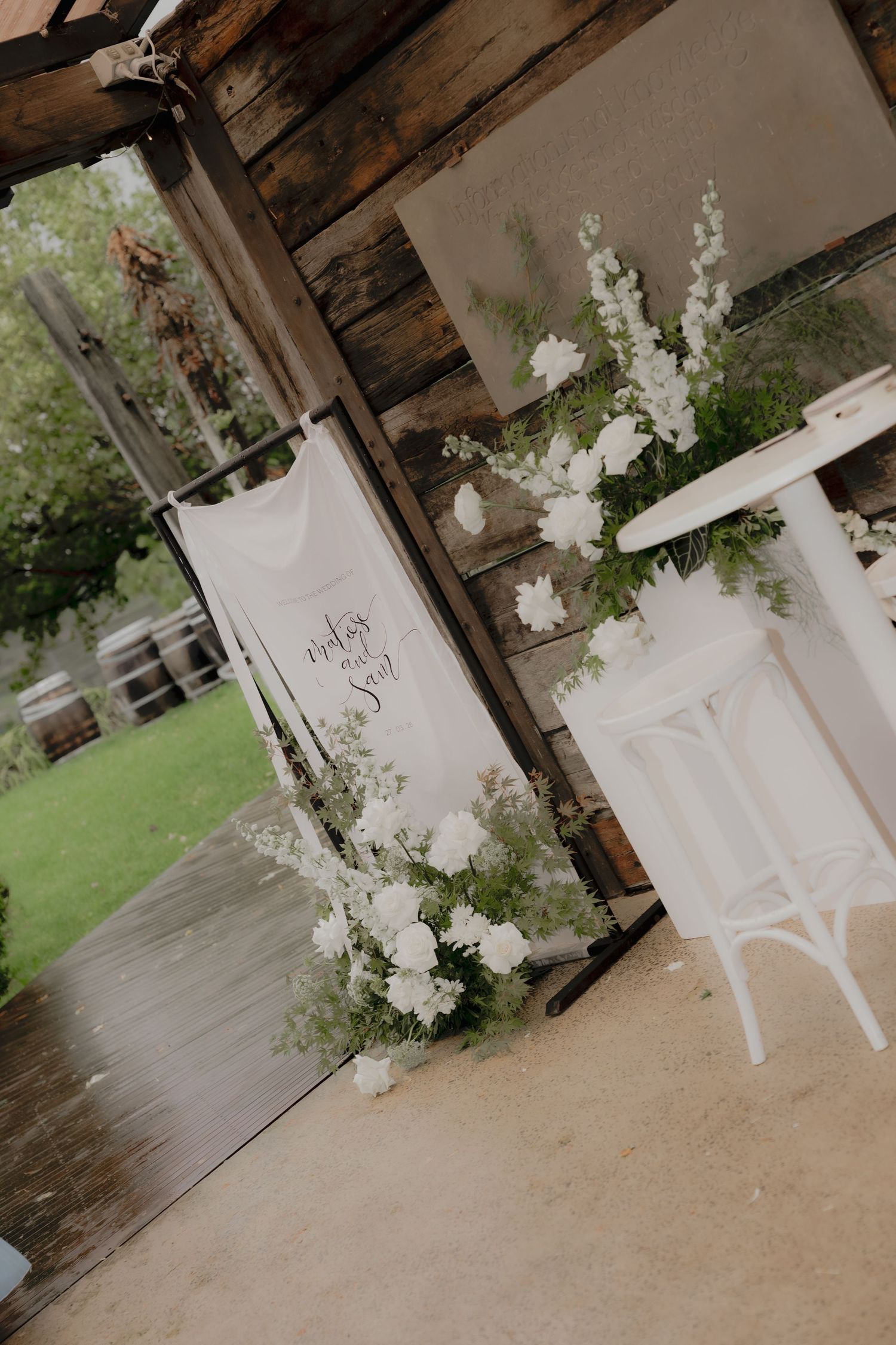 Elegant wedding venue with white floral arrangements, calligraphy banner, and white bistro table in rustic barn setting. Borrodell Vineyard