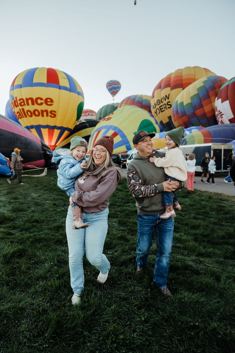Family of four smiling at a hot air balloon festival, holding two young children with colorful balloons inflating in background.