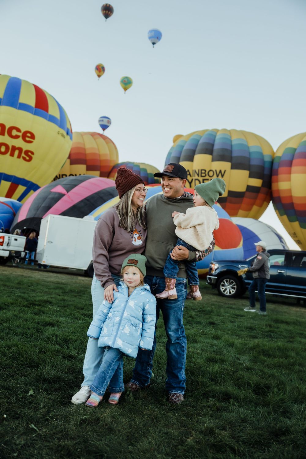 Family of four smiling at a hot air balloon festival with colorful balloons filling the sky behind them.