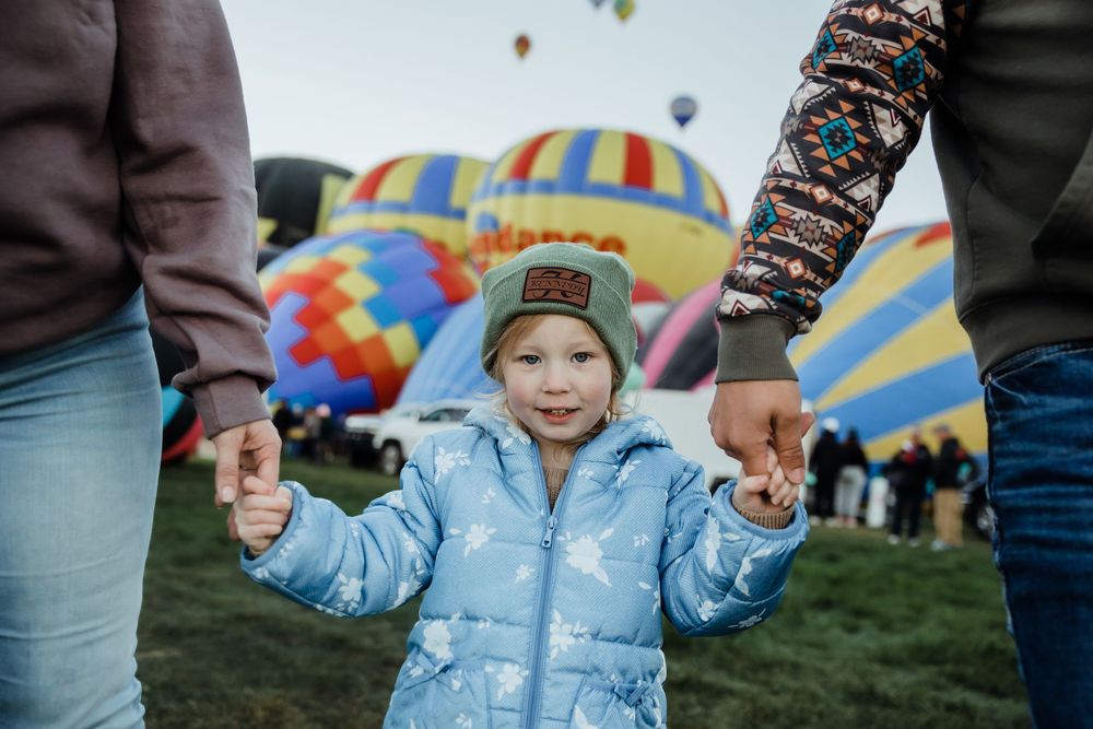 A toddler in a blue jacket holds hands with parents at a colorful hot air balloon festival.