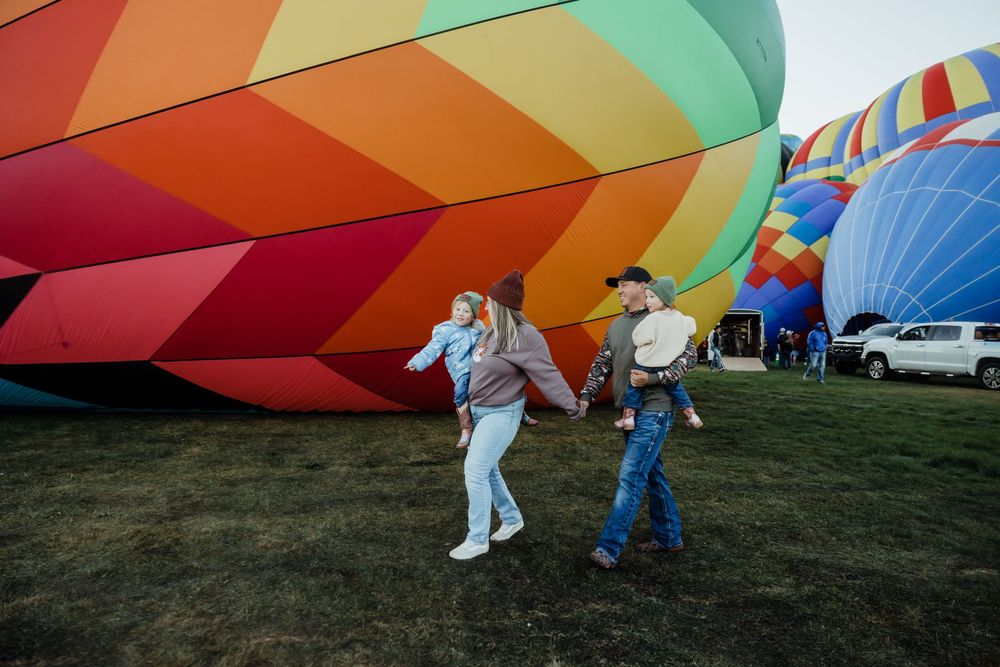 Family walking near colorful hot air balloons at an outdoor festival on a grassy field.
