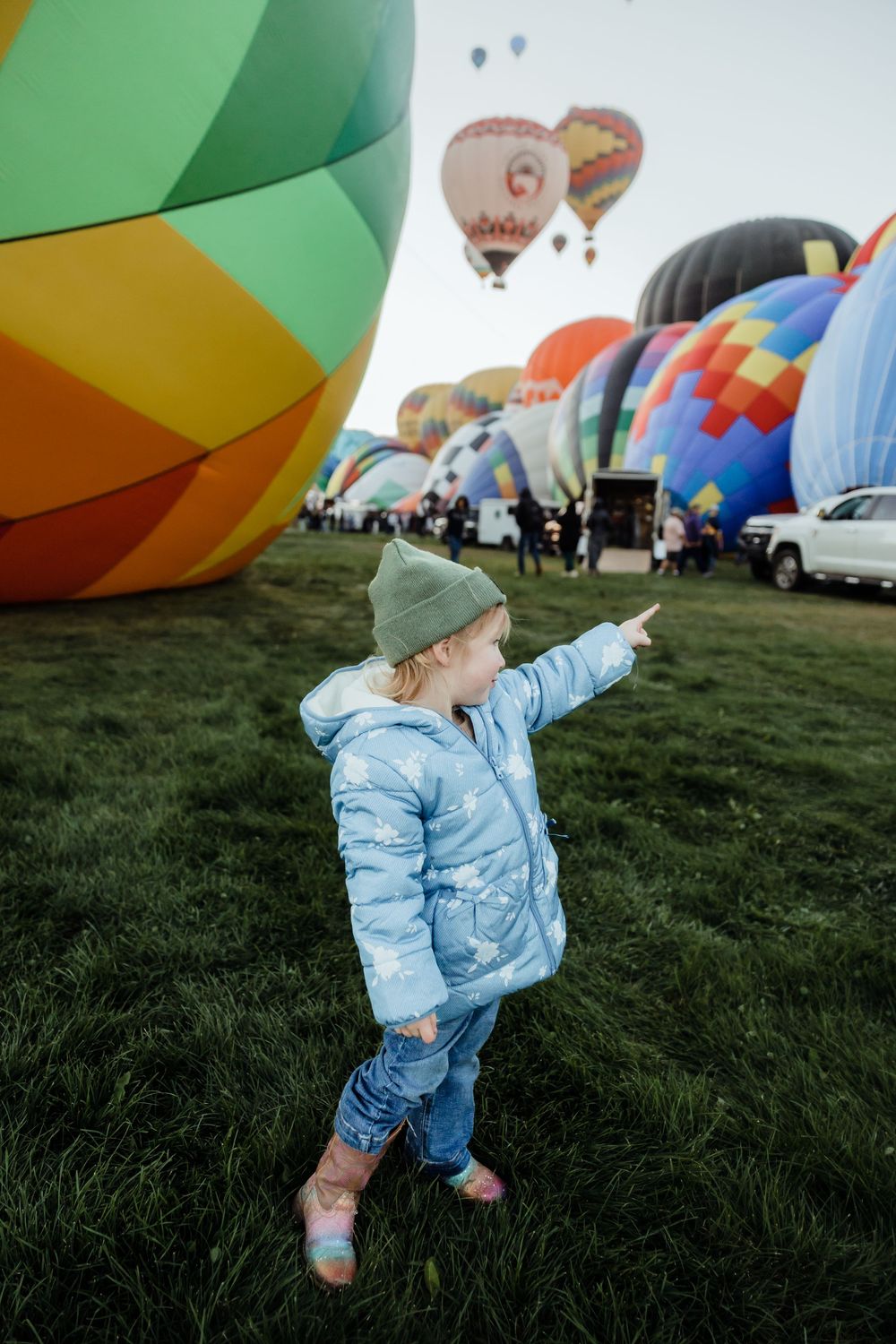 Toddler girl in blue jacket pointing at colorful hot air balloons at a festival on green grass.