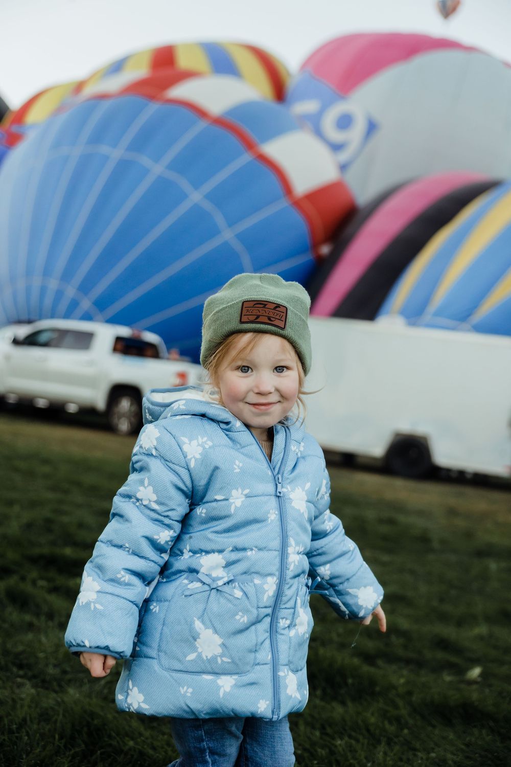 Toddler girl in blue puffer jacket and green hat smiles at hot air balloon festival.