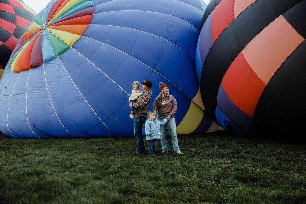 Family of four poses in front of colorful hot air balloons on green grass at an outdoor balloon festival.