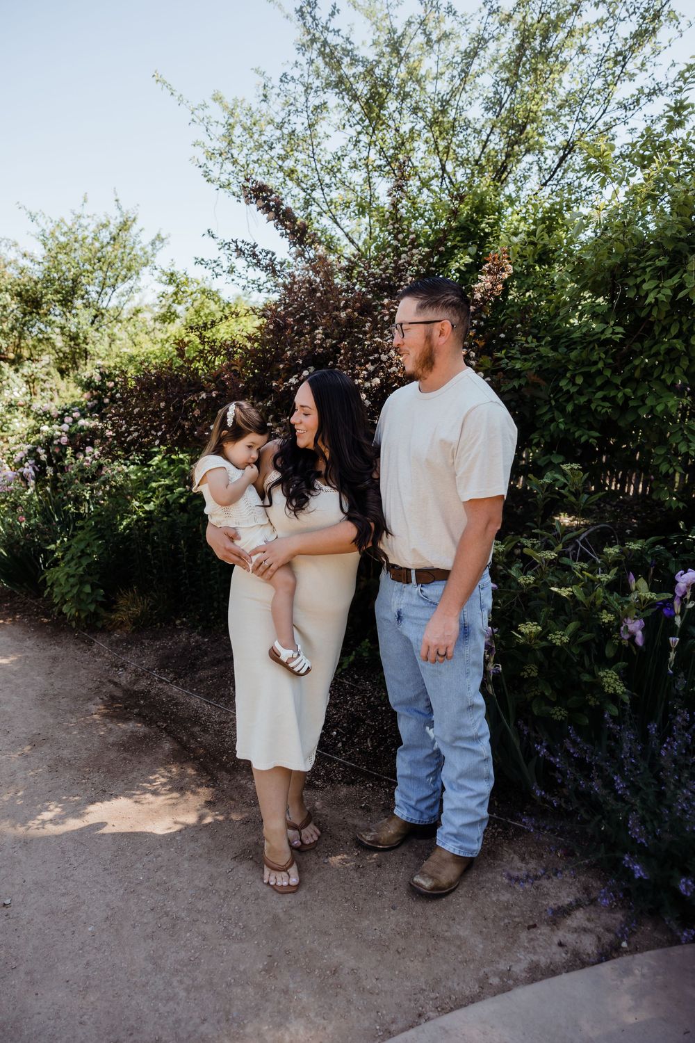 A young family poses outdoors, mother holding toddler in white dress, father in jeans and cream shirt, surrounded by lush greenery.