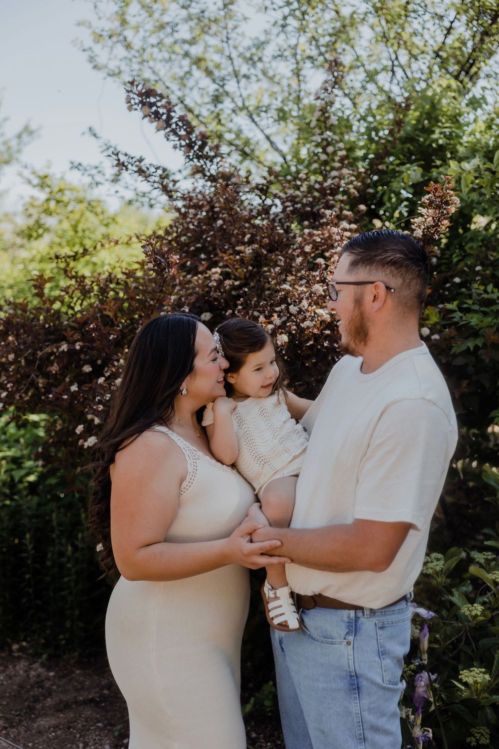 Pregnant woman in beige dress with partner holding toddler, posing outdoors among lush green trees and shrubs.