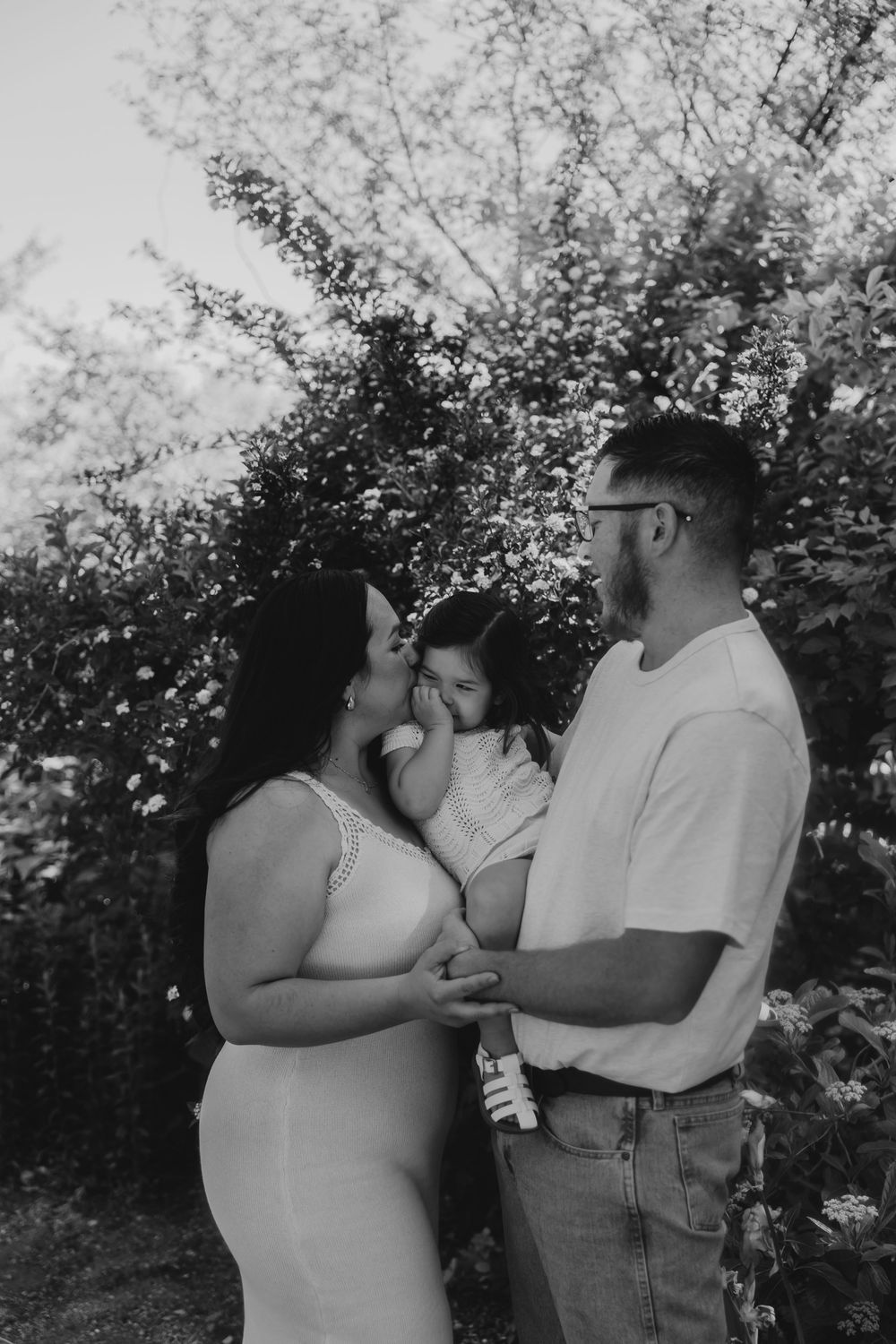 Black and white photo of a pregnant couple holding their toddler in a lush garden setting.