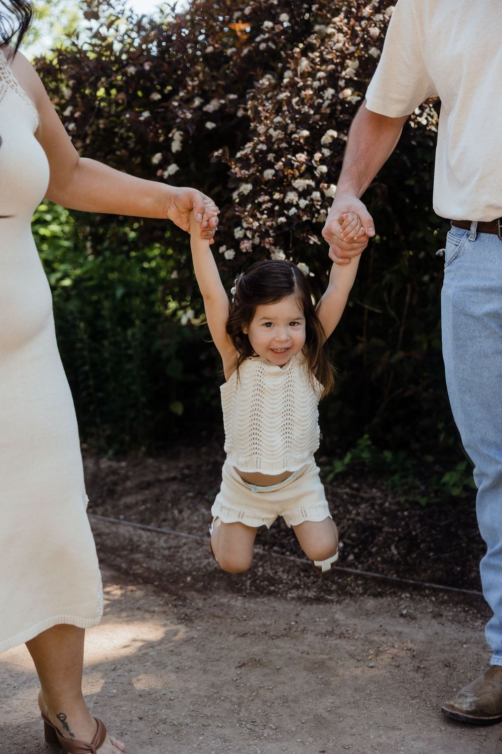 A joyful toddler girl in cream outfit is lifted by her parents hands outdoors near blooming trees.