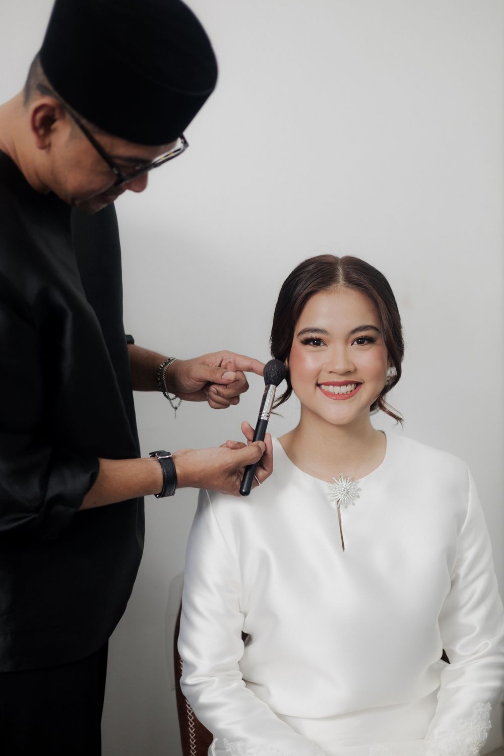 A makeup artist in black applies makeup to a smiling young woman in a white outfit against a light background.