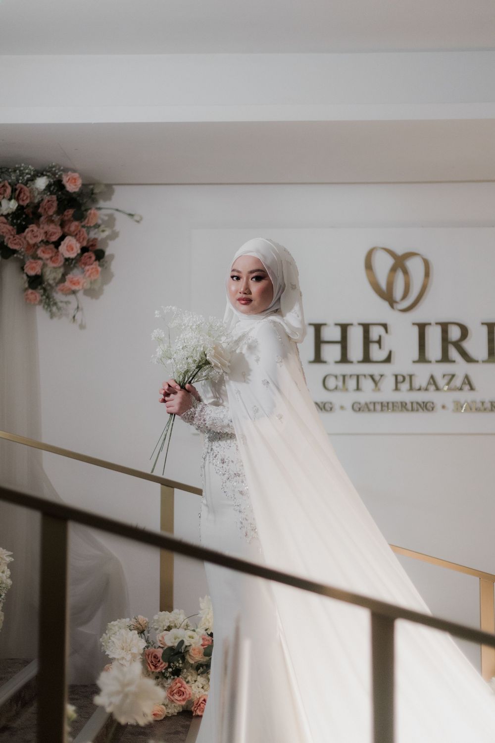 Muslim bride in white hijab and wedding gown holds bouquet at The Iris City Plaza wedding venue.