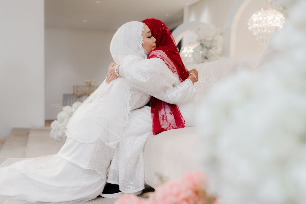 Two women in hijabs share a warm embrace at a wedding, one in white bridal attire and one in red.