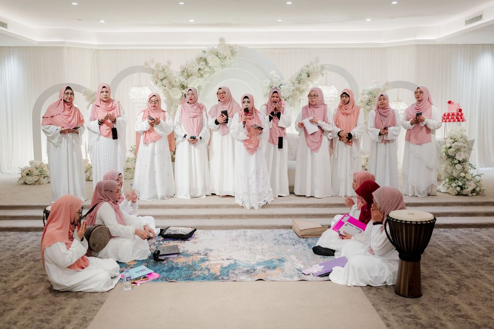 Muslim women in pink hijabs and white dresses gathered for a traditional wedding ceremony in an elegant decorated hall.