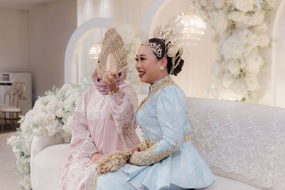 Two women in traditional Indonesian wedding attire sit together on a white sofa, laughing and holding a decorative fan.