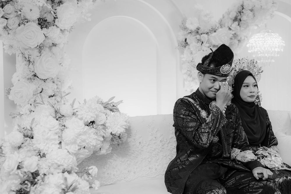 Black and white photo of a Malay couple in traditional attire sitting together at an elegant wedding ceremony.