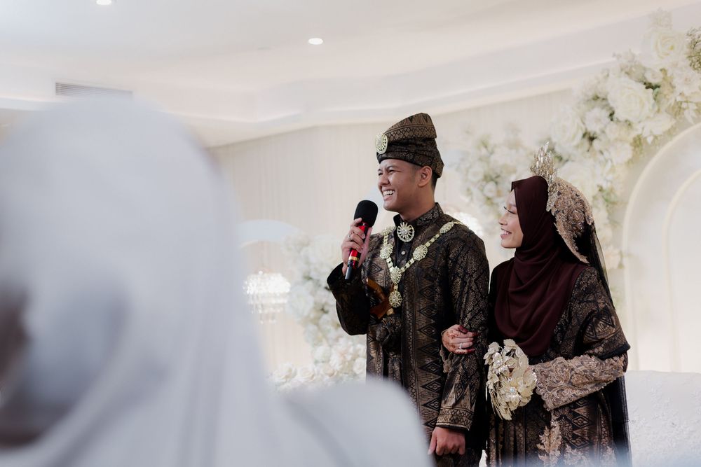 Couple in traditional Indonesian wedding attire smiling and speaking into a microphone at an elegant white floral ceremony.