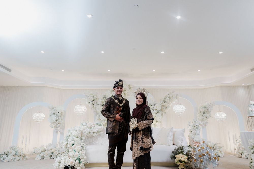 Couple in traditional Javanese wedding attire posing in an elegant white floral decorated reception hall.