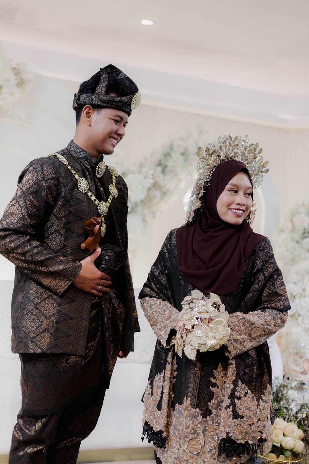 A couple in traditional Malay wedding attire, wearing black and gold outfits, with the bride holding a white bouquet.