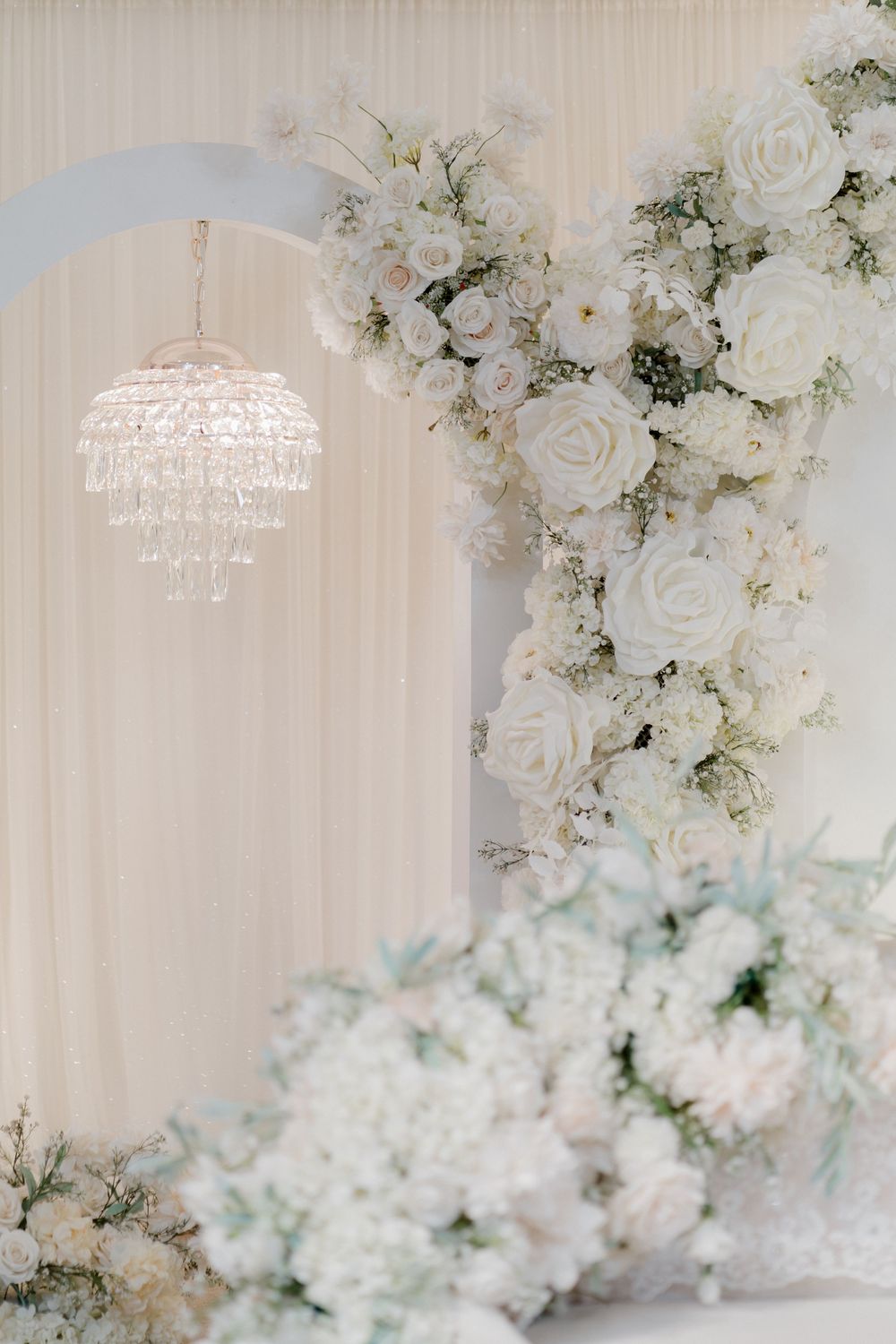 Elegant wedding backdrop with white roses, crystal chandelier, and lush floral arrangements in soft ivory tones.