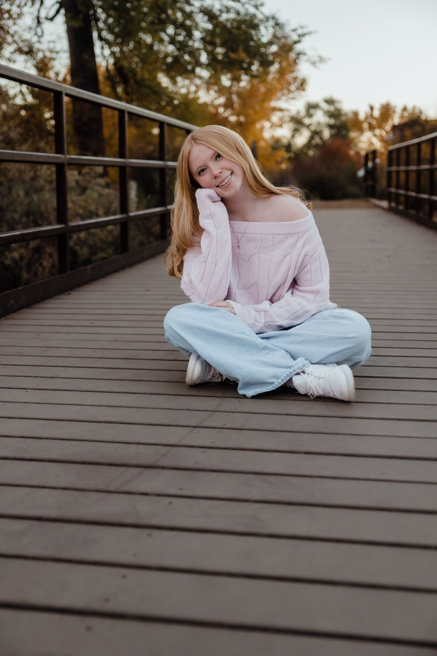 Smiling blonde girl sitting cross-legged on a wooden bridge in a pink off-shoulder top and light blue jeans.
