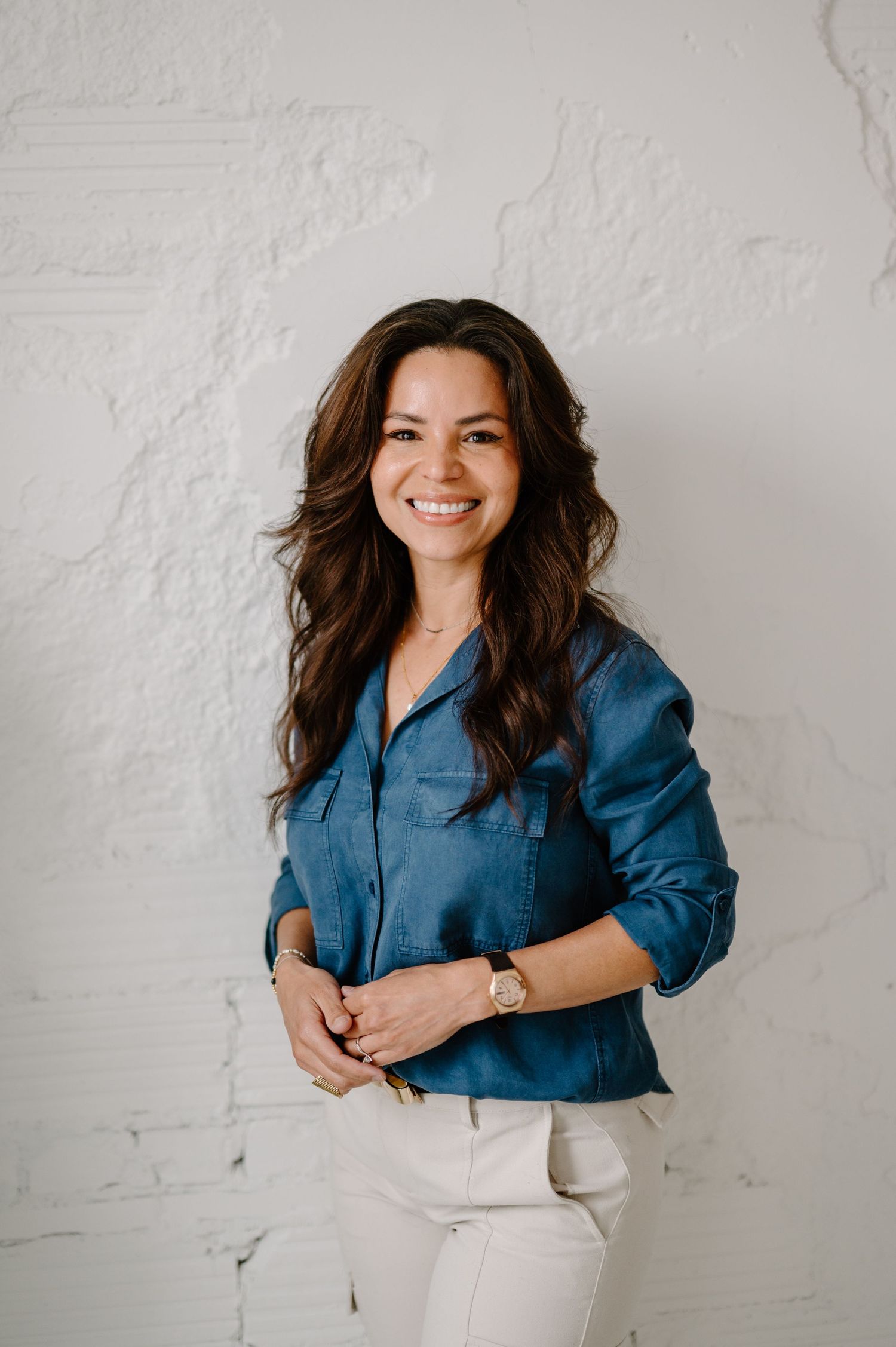 Smiling woman with long brown hair wearing a blue denim shirt and white pants, posing against a white brick wall.