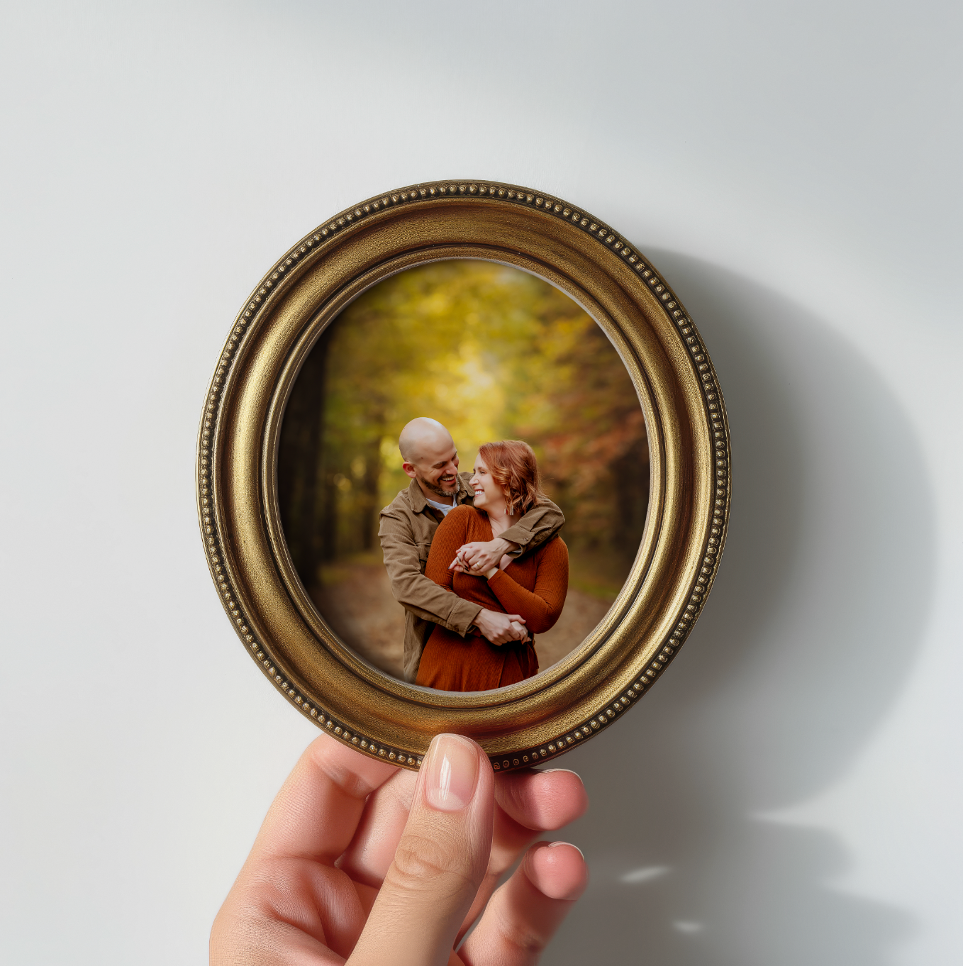 Hand holding a small oval gold frame with a photo of an elderly couple embracing in an autumn forest setting.