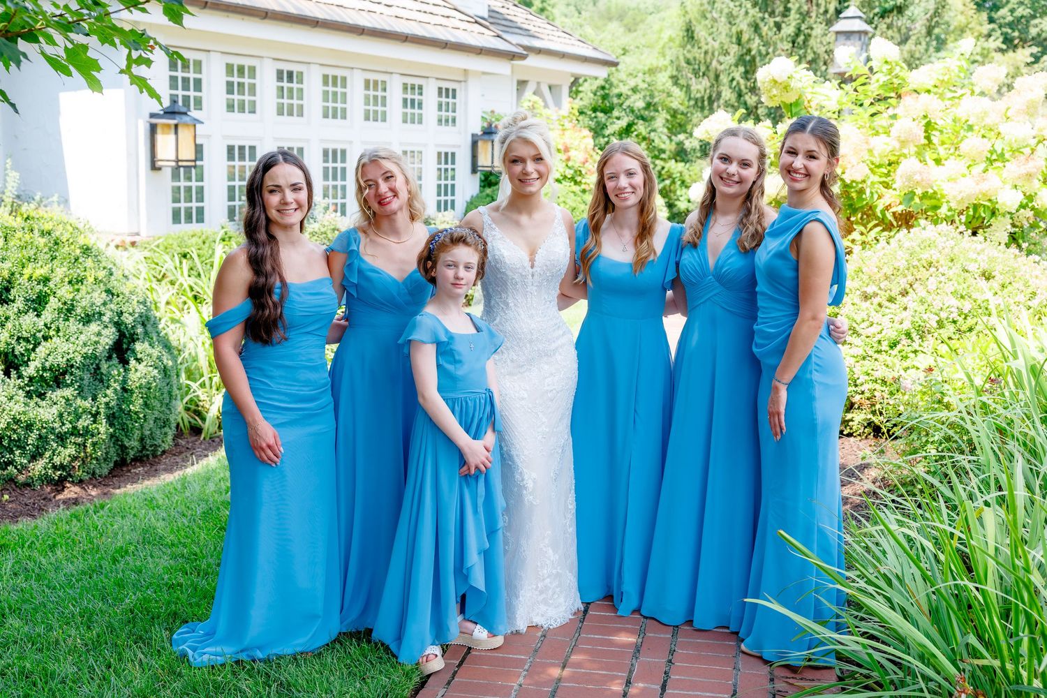 Bride in white lace gown poses with bridesmaids in blue dresses and flower girl on brick path outside elegant venue.