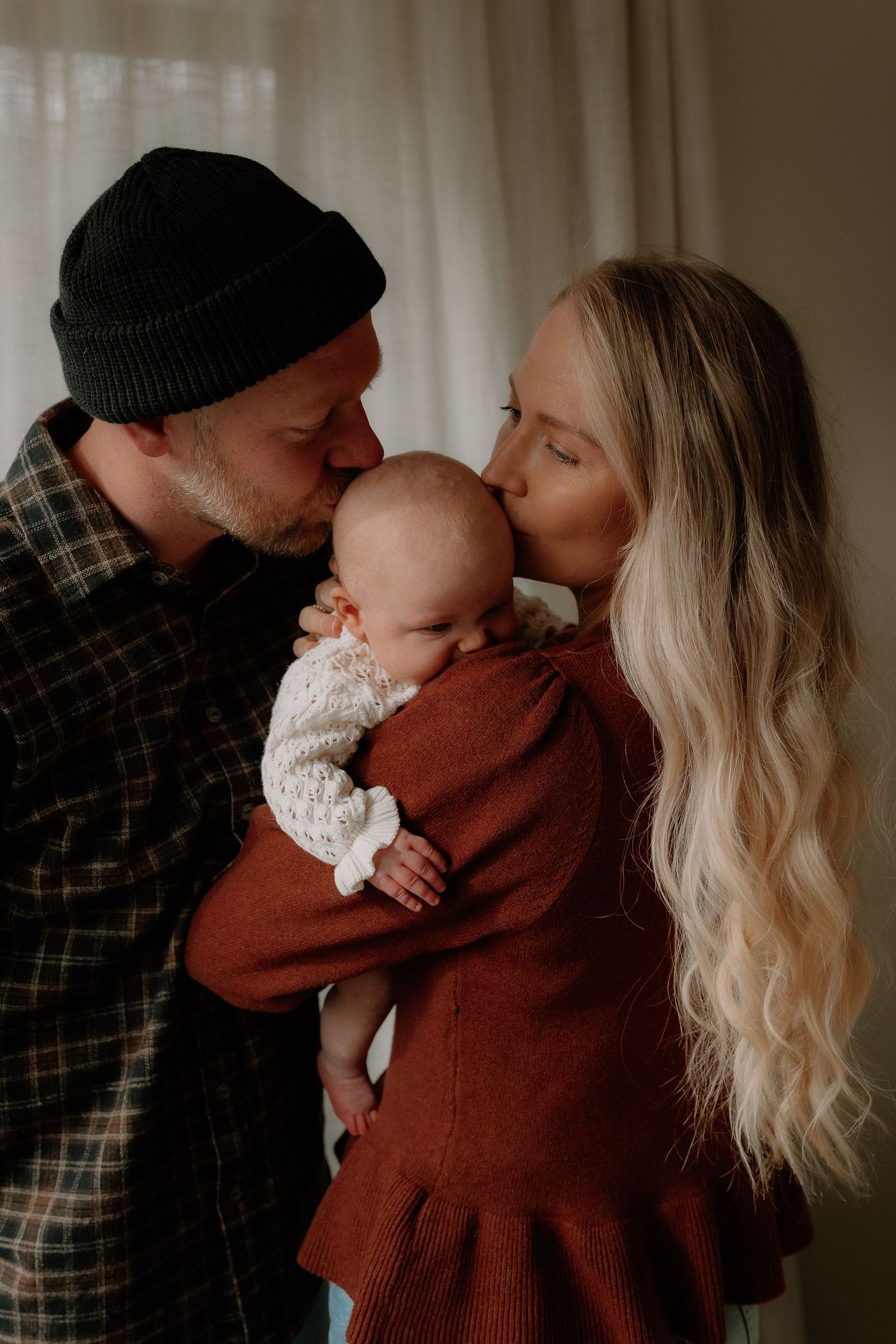 A couple tenderly kisses their newborn baby, the mother in a rust dress and father in a plaid shirt and beanie.