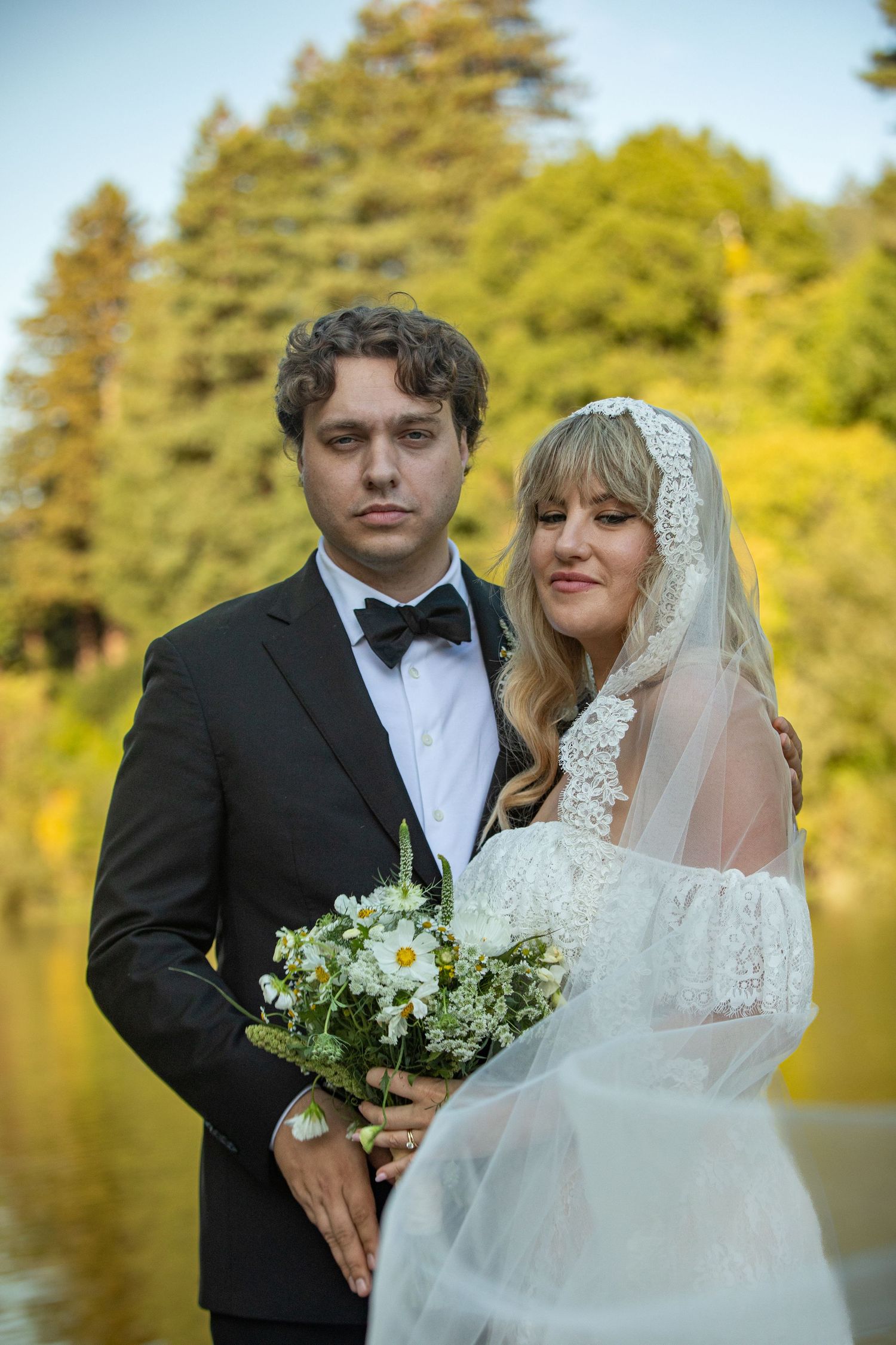Bride and groom pose outdoors near a lake, she holds a daisy bouquet in a lace veil, he wears a black tuxedo.