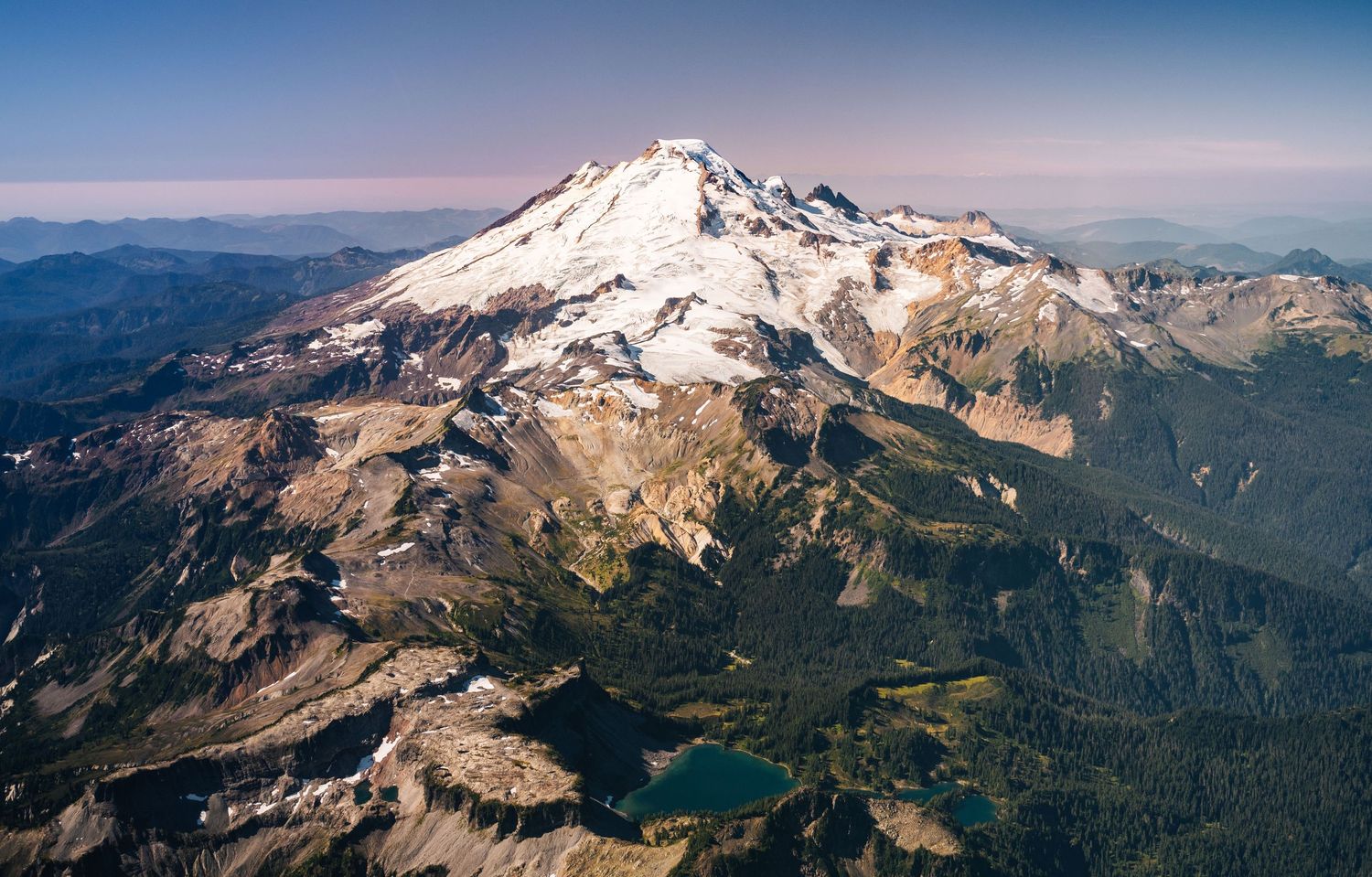 Aerial view of snow-capped Mount Baker in Washington state with rugged mountain terrain and wilderness stretching to horizon.