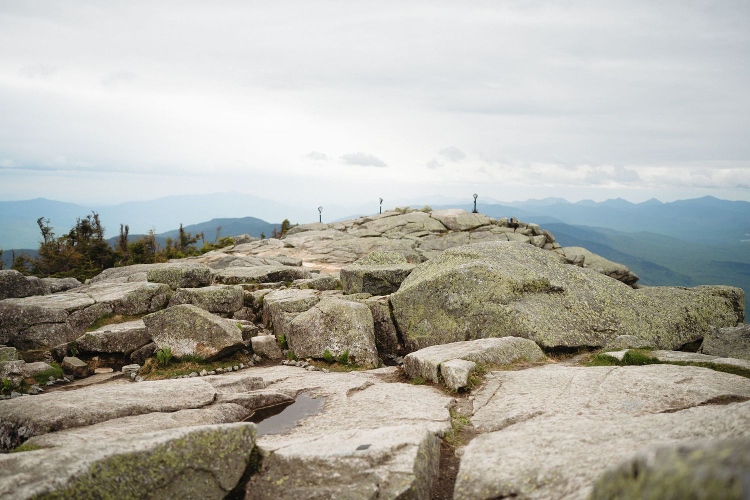 Rocky mountain summit with expansive views of green valleys and misty peaks under an overcast sky.