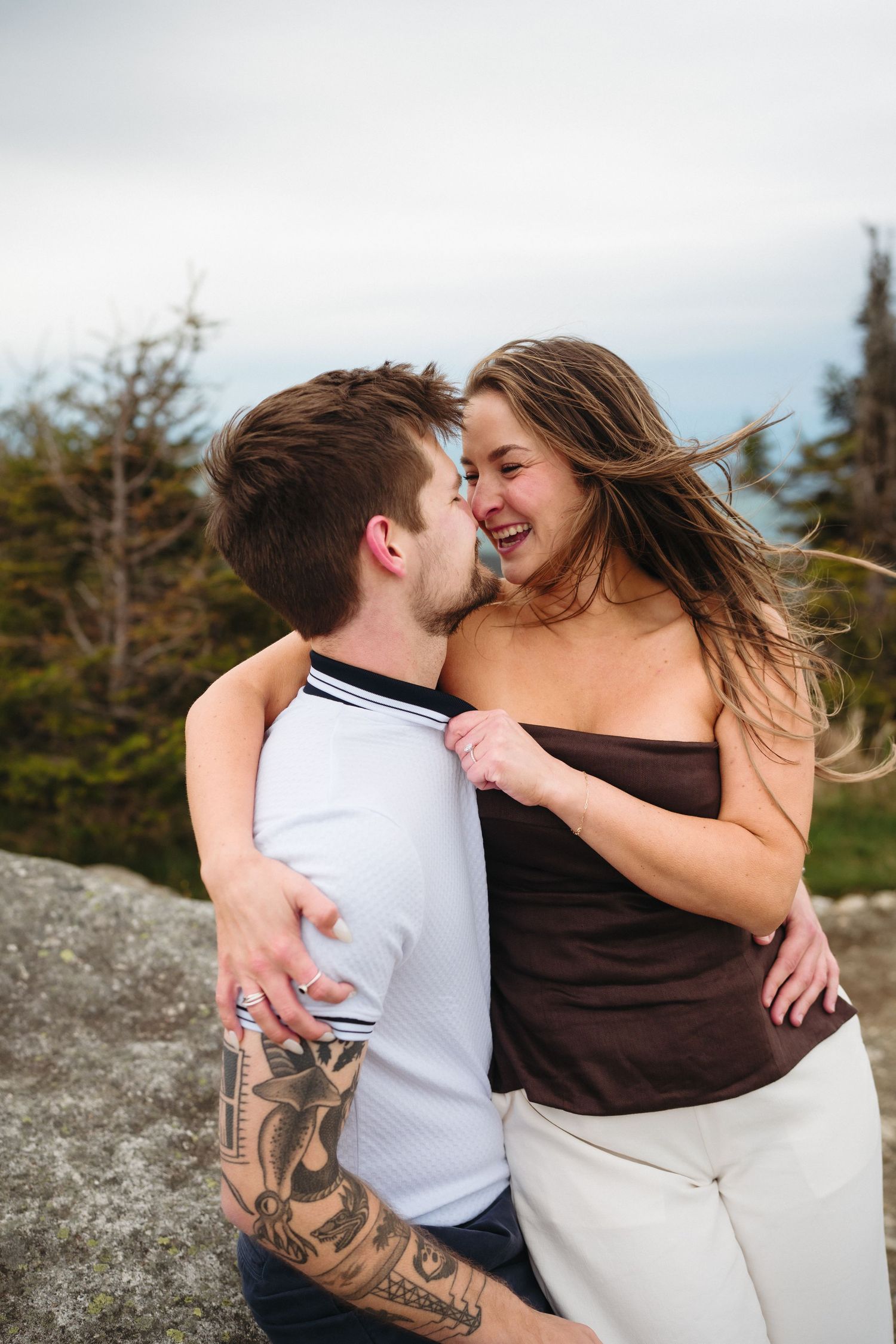 A couple embraces and laughs together outdoors on a rocky overlook, the man showing tattoos on his arm.