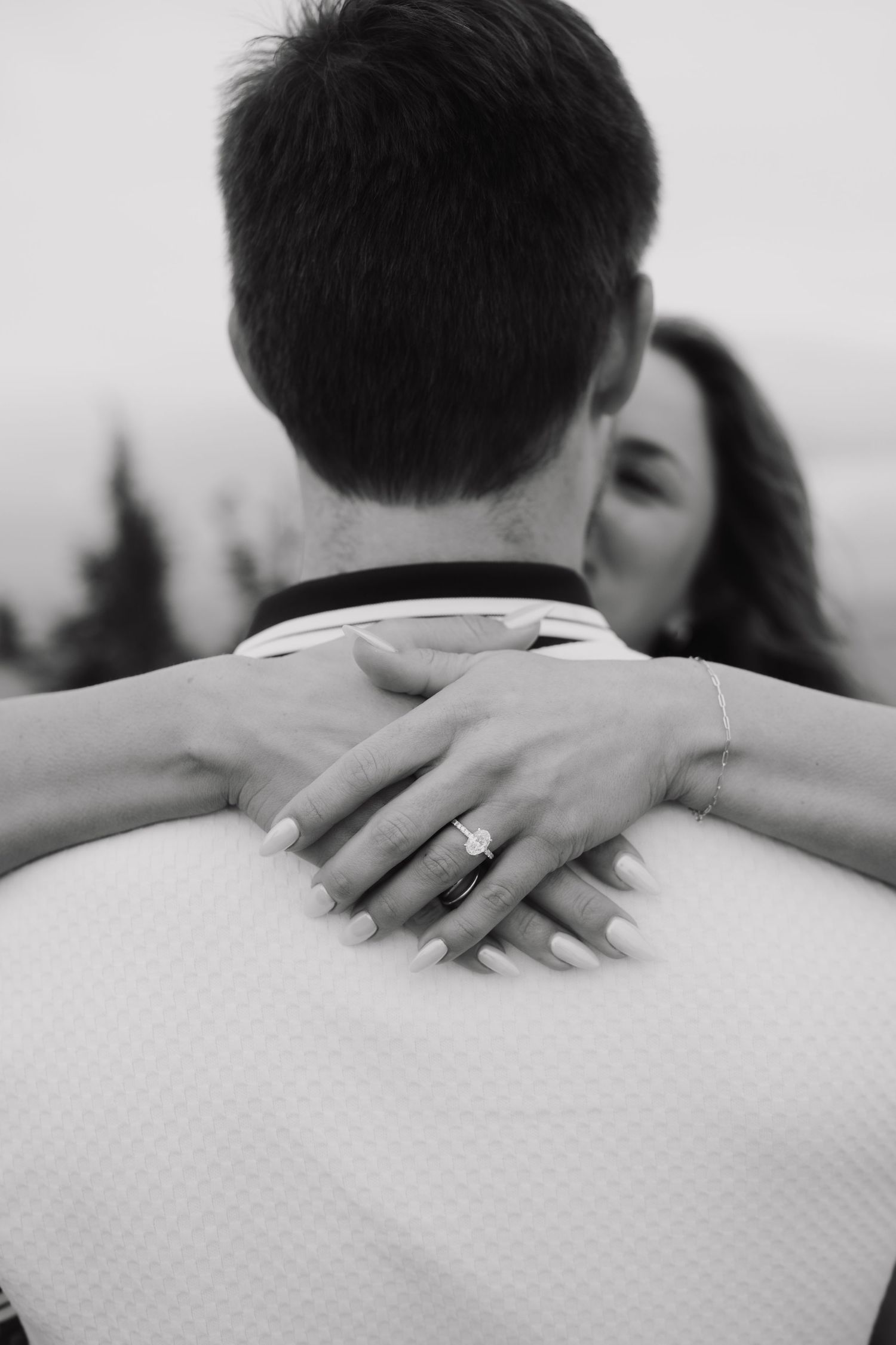 Black and white photo of couple embracing, woman showing engagement ring while kissing man from behind.