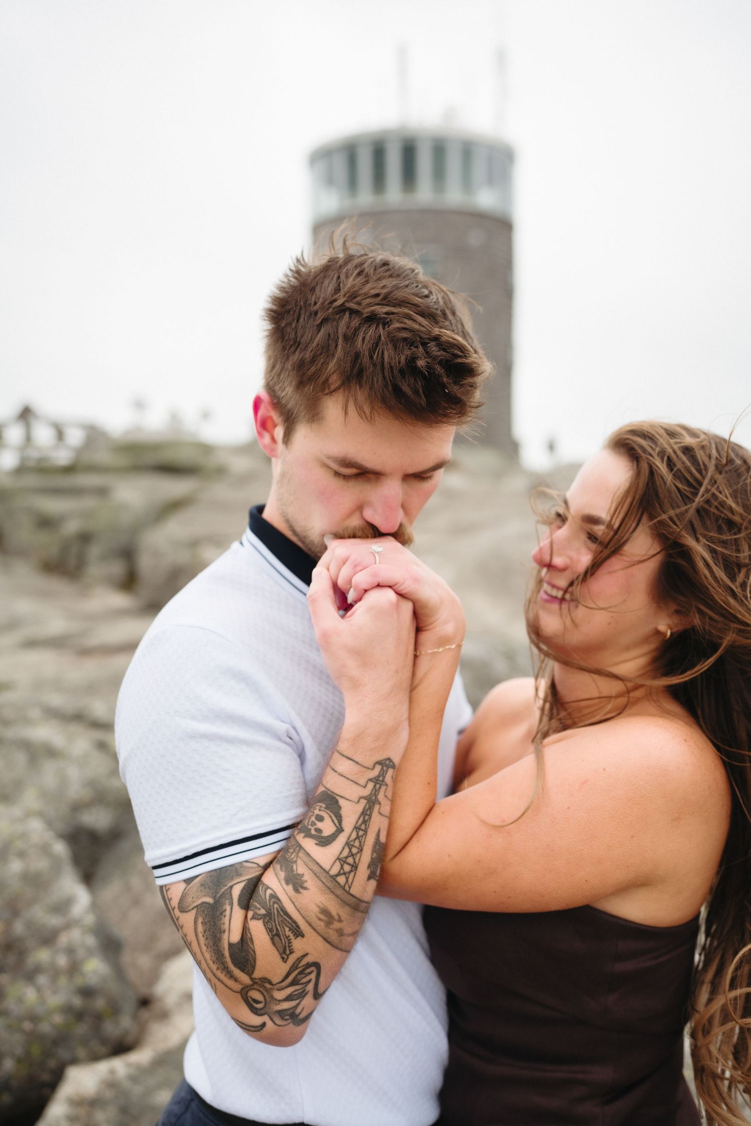 Tattooed man kissing womans hand near stone tower on mountain summit.