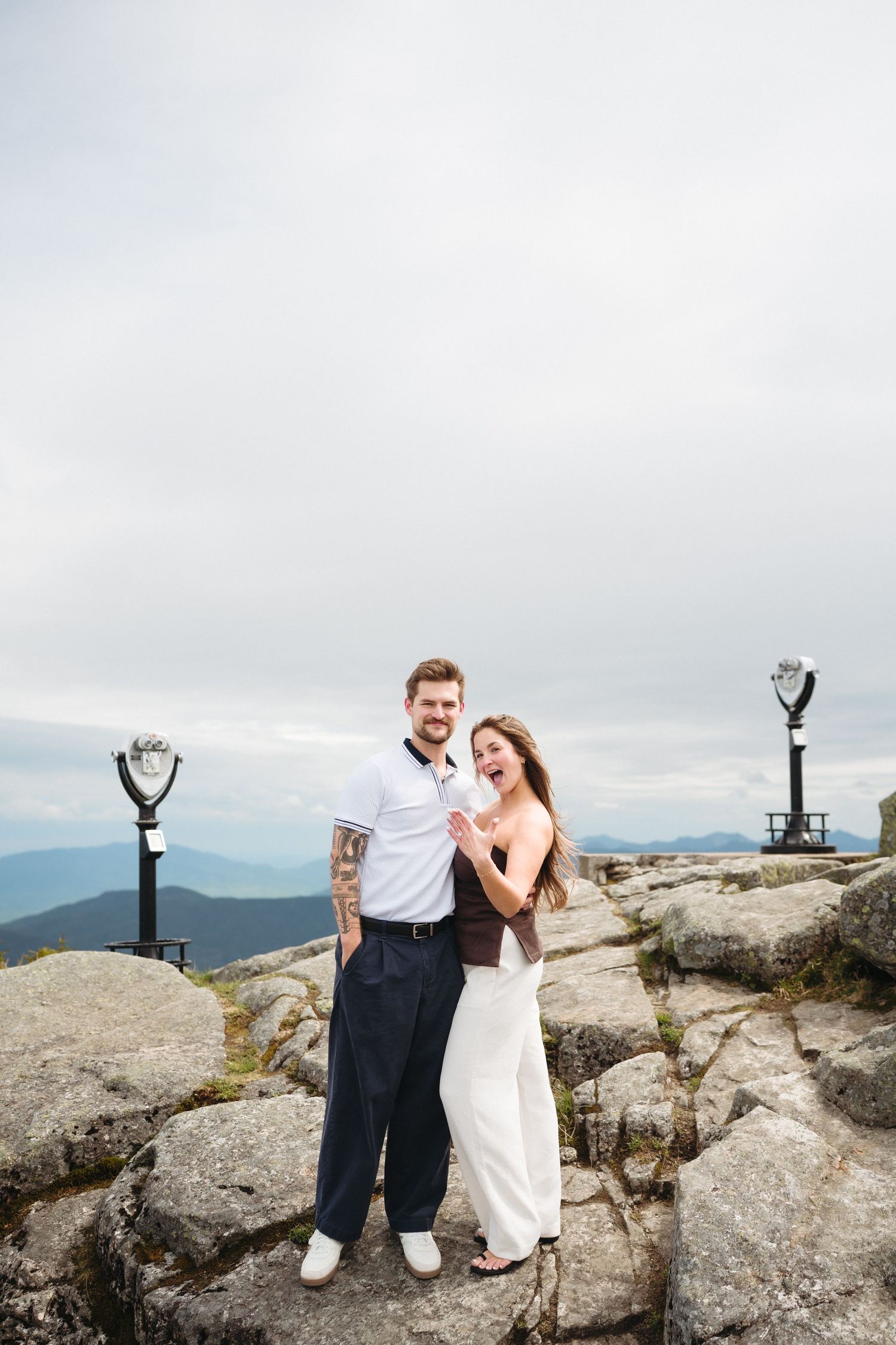 Happy couple posing on rocky mountain top with panoramic views and coin-operated binoculars nearby.