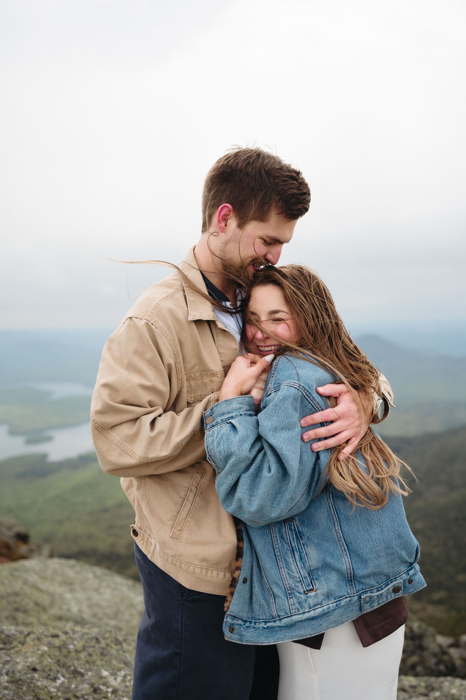 Couple embracing on mountain summit, man in tan jacket hugging smiling woman in denim jacket with scenic valley view.