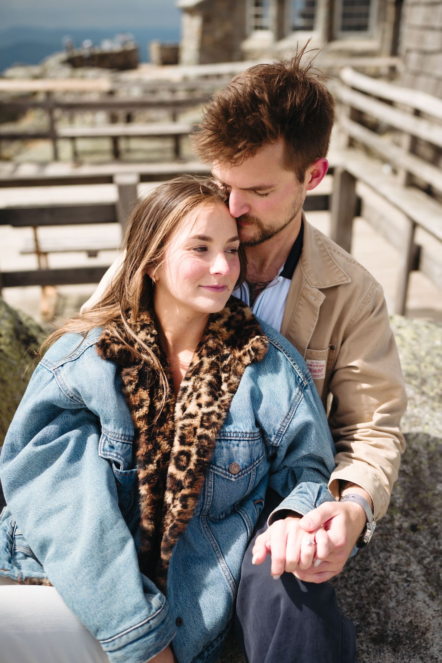 A couple shares a tender moment outdoors, woman in denim and leopard print jacket, man in tan jacket kissing her forehead.