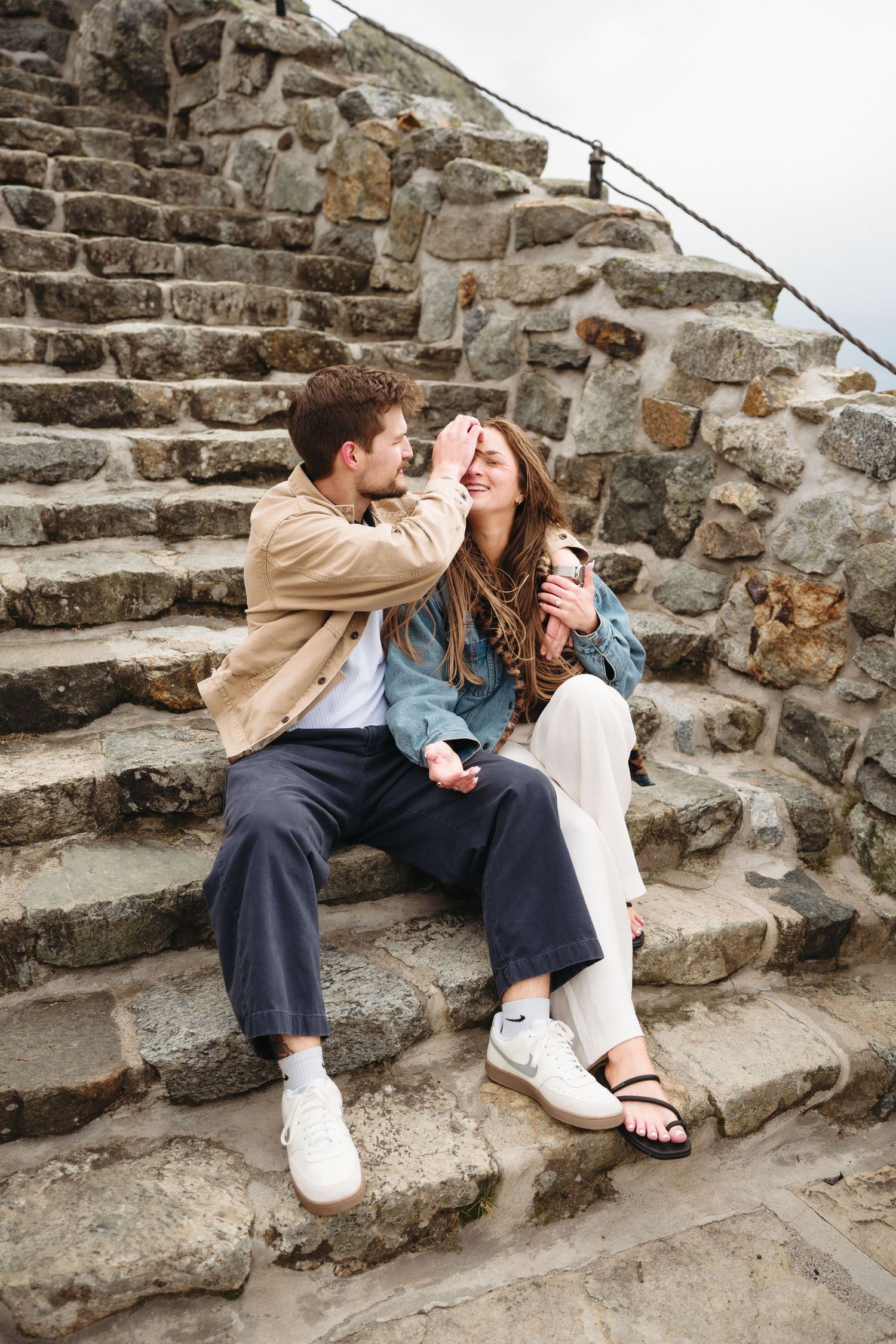 Playful couple sitting on rustic stone steps laughing and touching faces.