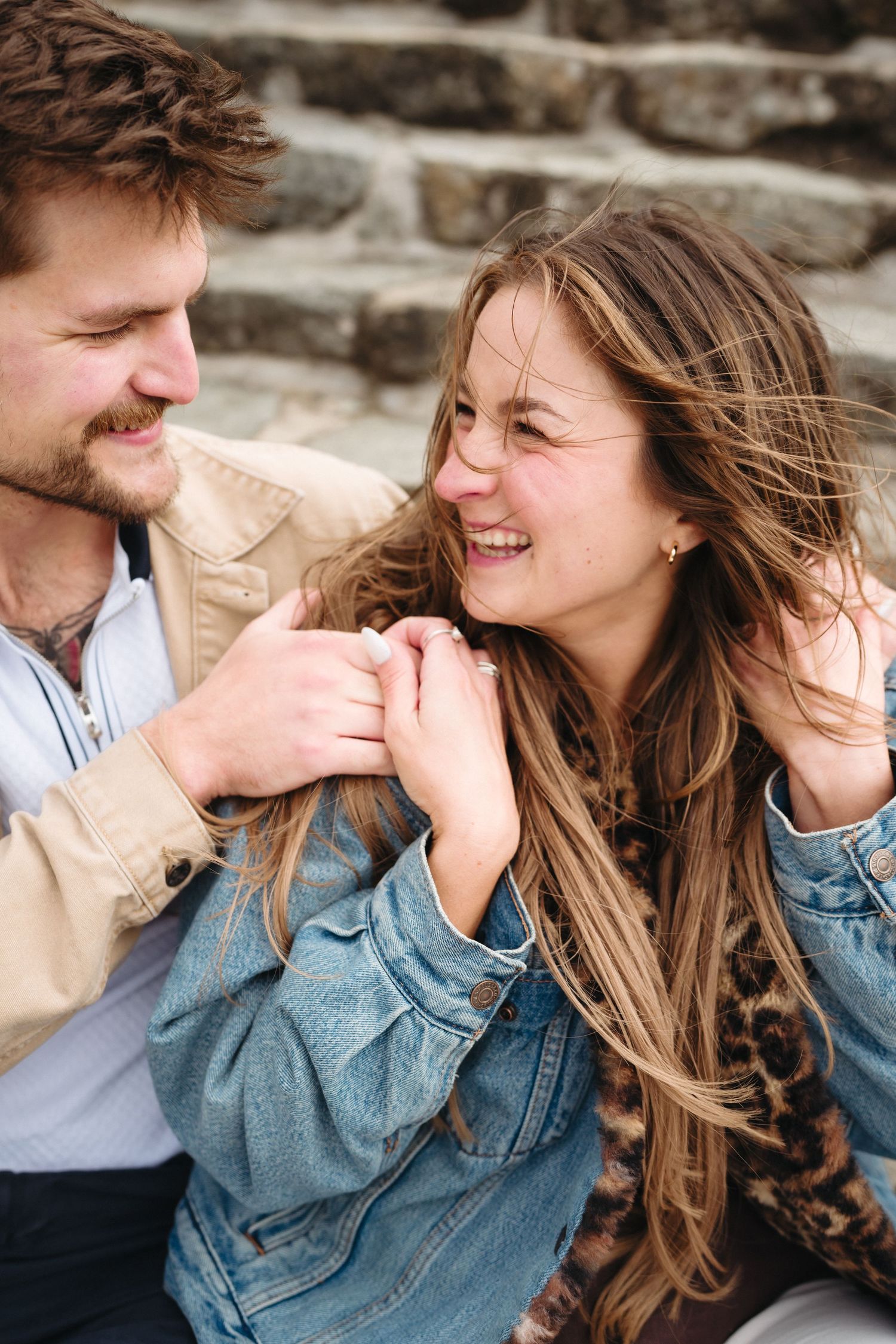 Happy couple sitting on stone steps, smiling at each other while holding hands, wearing denim jackets outdoors.