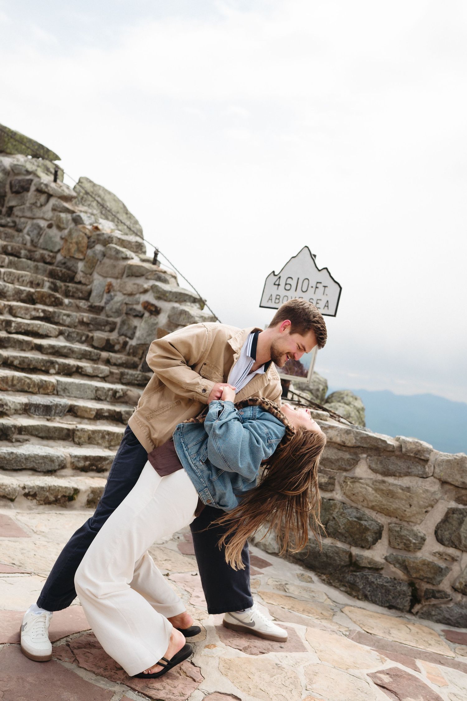 Couple dancing romantically at a mountain summit sign reading 4610ft above sea level on a stone overlook.