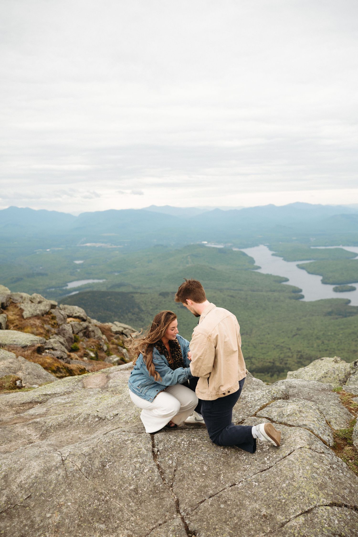 Couple sharing a romantic proposal moment on a rocky mountain summit with scenic valley and lake views below.