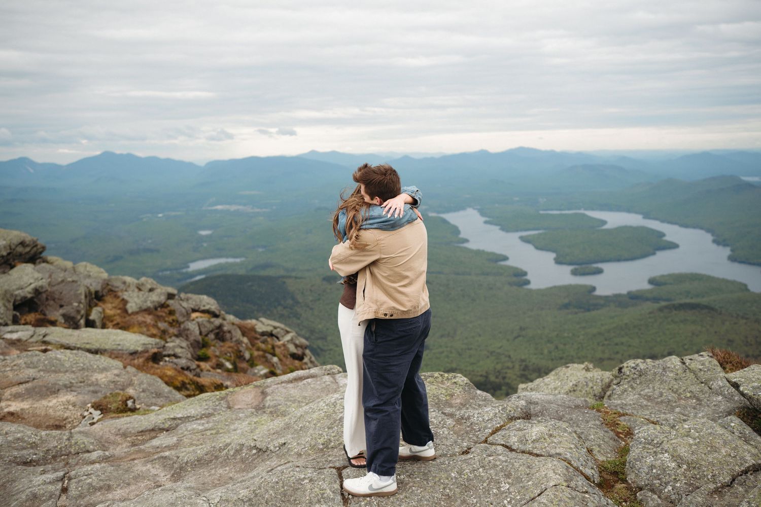 Couple embracing on rocky mountain summit with panoramic views of lakes and green forest valleys below.