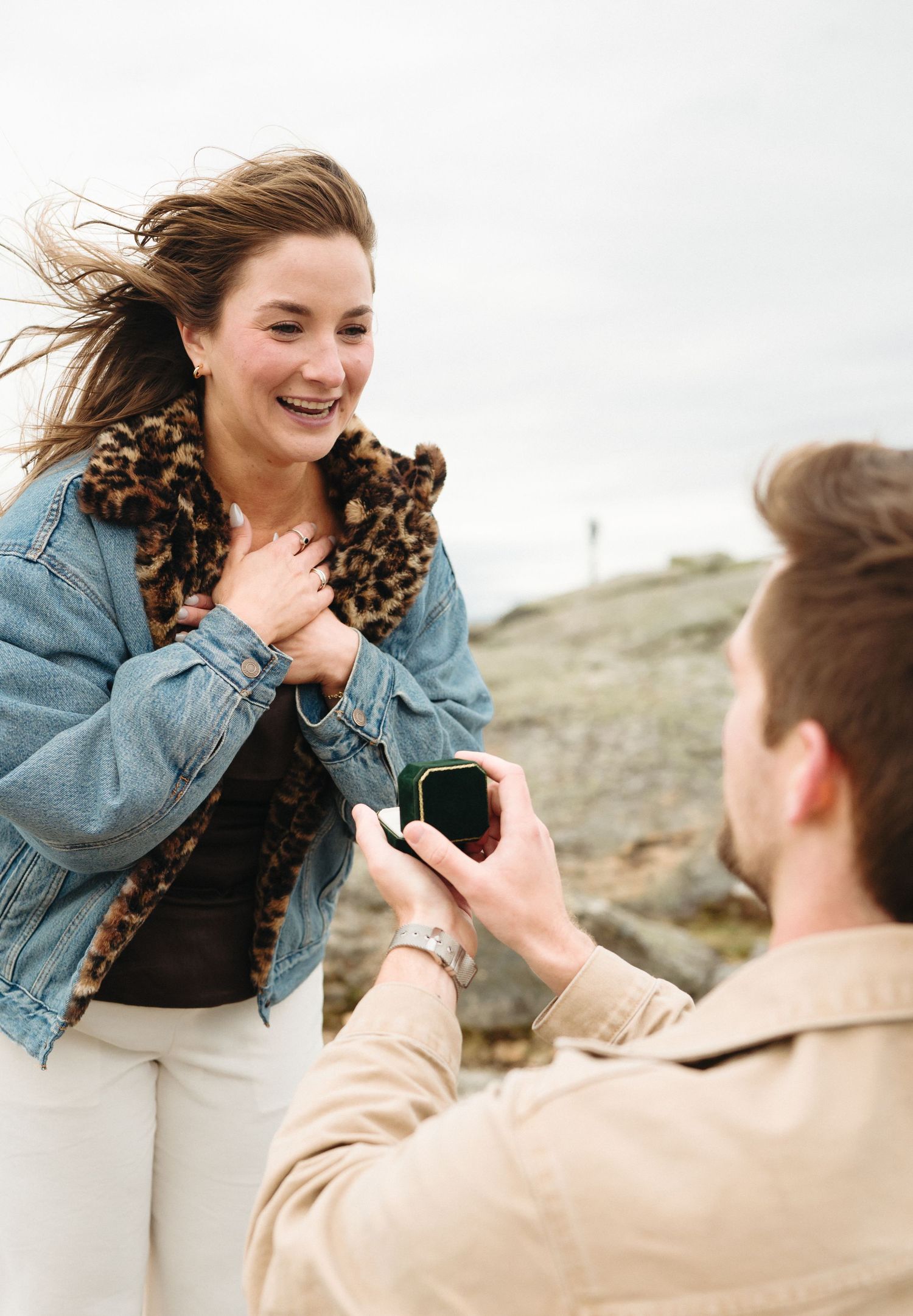 Man kneeling outdoors proposing with a ring box to a smiling woman in a denim and leopard print jacket.